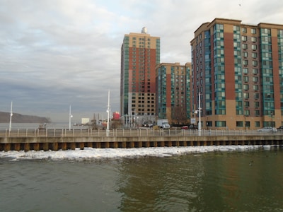 High-rise buildings with blue and red accents stand adjacent to a body of water. The waterfront area has a promenade with railings, and the water appears calm with a few visible boats and sparse traffic on the road.