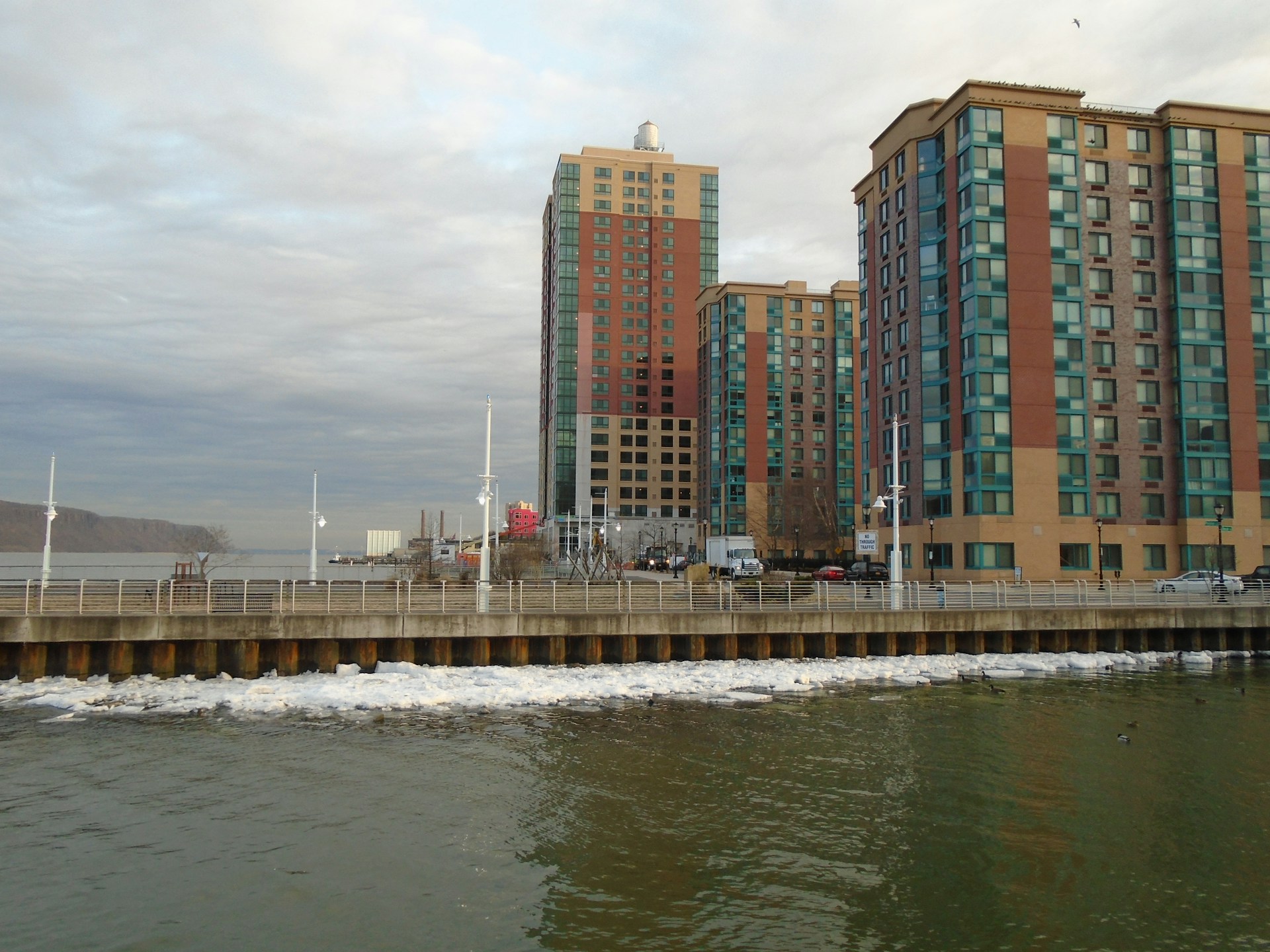 High-rise buildings with blue and red accents stand adjacent to a body of water. The waterfront area has a promenade with railings, and the water appears calm with a few visible boats and sparse traffic on the road.