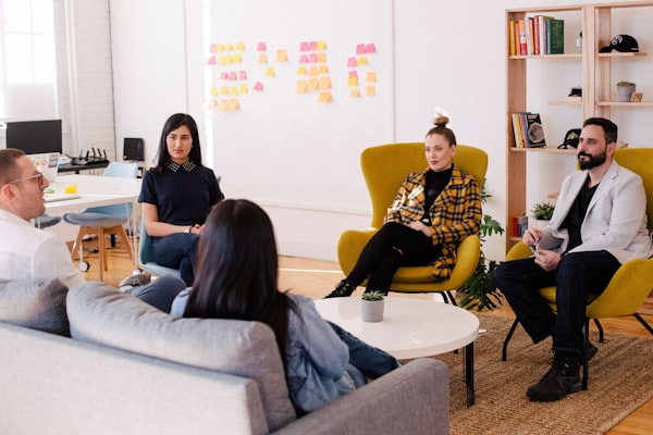 Trois femmes et deux hommes assis sur des fauteuils et un canapé discutant devant un tableau avec des post-its