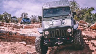 A rugged Jeep navigating a rocky, off-road terrain with large tires gripping the uneven surface. Two more Jeeps are visible in the background, also maneuvering through the challenging landscape. The scene is set in a desert-like environment with scattered shrubs and a cloudy sky overhead.