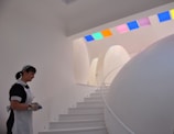 A futuristic interior setting features smooth, egg-shaped structures and a curved white staircase with small blue lights embedded in each step. A person wearing a maid's uniform is present on the left side, holding an object. The ceiling is adorned with colorful, square glass panels that cast colored light onto the walls.