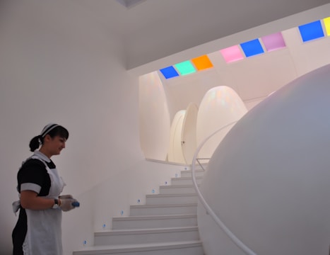 A futuristic interior setting features smooth, egg-shaped structures and a curved white staircase with small blue lights embedded in each step. A person wearing a maid's uniform is present on the left side, holding an object. The ceiling is adorned with colorful, square glass panels that cast colored light onto the walls.