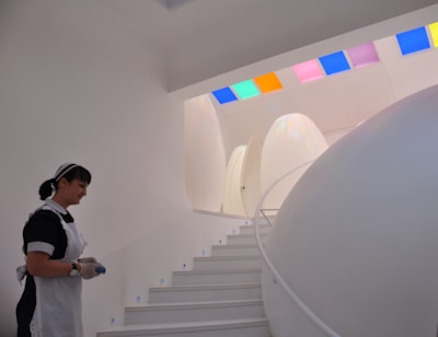 A futuristic interior setting features smooth, egg-shaped structures and a curved white staircase with small blue lights embedded in each step. A person wearing a maid's uniform is present on the left side, holding an object. The ceiling is adorned with colorful, square glass panels that cast colored light onto the walls.
