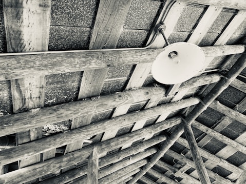 The image features an overhead view of a wooden ceiling structure with exposed beams. The beams are constructed from rough, unfinished wood, showcasing a rustic design. There is a single round, white light fixture attached to the beams, though the bulb appears to be missing or not illuminated. The space between the beams reveals a textured surface above.