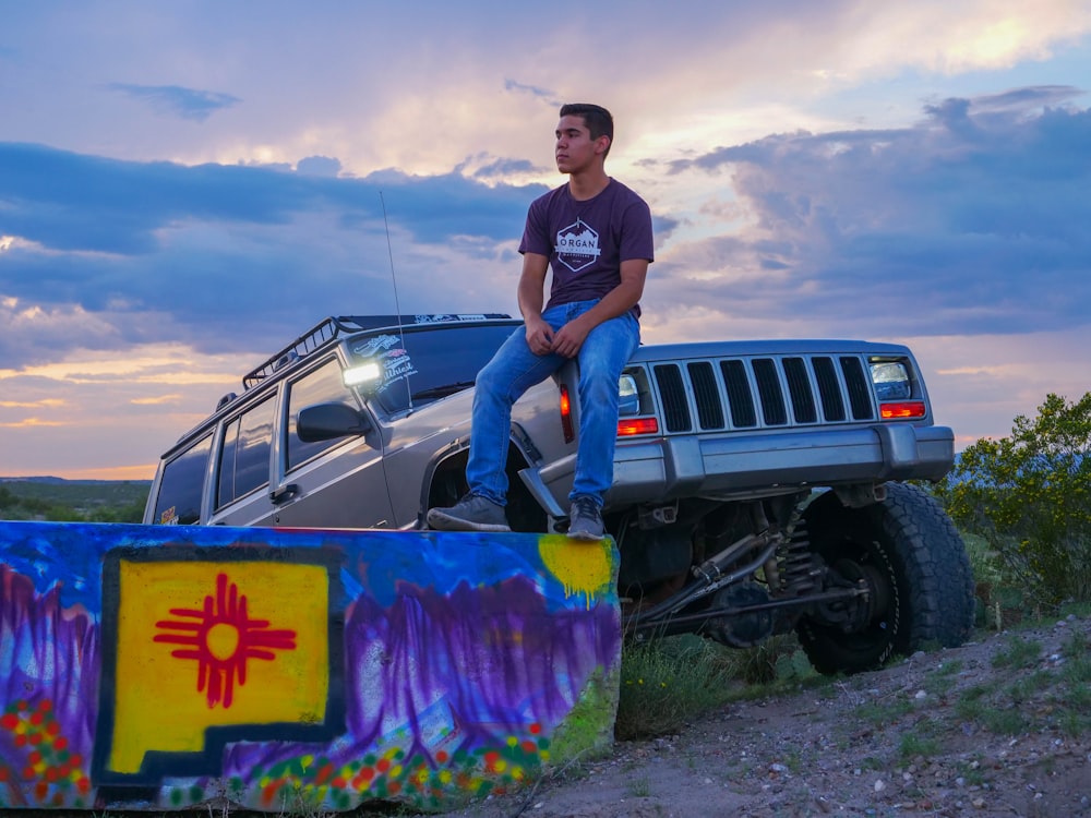 Mann sitzt auf Motorhaube eines geparkten grauen Jeep Cherokee SUV