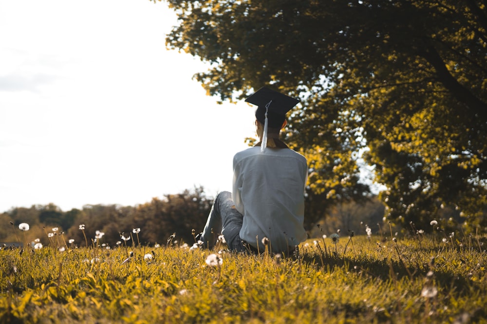 person in white shirt sitting on green grass field with black mortar cap on head