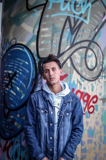 A young man leaning against a graffiti wall, proudly wearing a sovereign and sons hoodie.