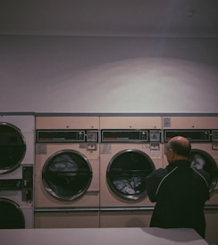 A person stands in front of a row of industrial-grade dryers in a dimly lit laundromat. The machines are beige with dark-tinted glass, and the one in use shows clothes tumbling inside. The walls are plain and the environment gives an impression of quiet solitude.