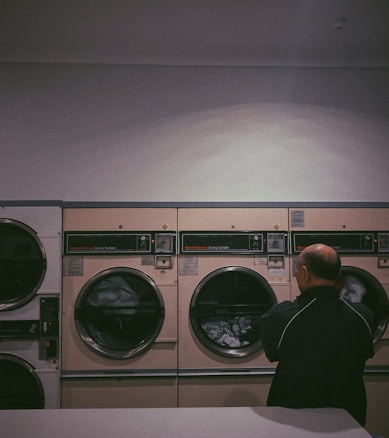A person stands in front of a row of industrial-grade dryers in a dimly lit laundromat. The machines are beige with dark-tinted glass, and the one in use shows clothes tumbling inside. The walls are plain and the environment gives an impression of quiet solitude.