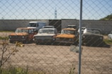 Several vintage cars are parked in a line behind a chain-link fence in a dusty, open lot. A few plants and shrubs are visible in the foreground with a brick wall serving as a backdrop. The cars appear aged and possibly abandoned.