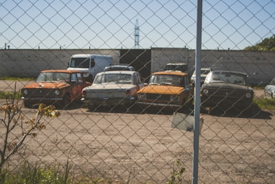Several vintage cars are parked in a line behind a chain-link fence in a dusty, open lot. A few plants and shrubs are visible in the foreground with a brick wall serving as a backdrop. The cars appear aged and possibly abandoned.