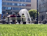 A group of people sits on a bench in front of a fountain, with water jets arching gracefully. The scene takes place in an urban setting with a large building in the background. The lush green grass in the foreground and trees suggest a park-like environment.