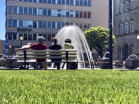 A group of people sits on a bench in front of a fountain, with water jets arching gracefully. The scene takes place in an urban setting with a large building in the background. The lush green grass in the foreground and trees suggest a park-like environment.
