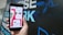 A hand holding a smartphone displaying a mobile app interface with a 'Match' notification. The screen shows profile pictures and chat options in a colorful design. The background is blurred with large text in blue and white.