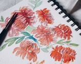 A watercolor painting of vibrant orange-red flowers with green leaves on a spiral-bound sketchbook. A black paintbrush rests on the paper, indicating the artistic process.
