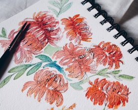 A watercolor painting of vibrant orange-red flowers with green leaves on a spiral-bound sketchbook. A black paintbrush rests on the paper, indicating the artistic process.