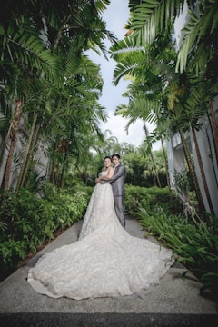 A couple stands embraced in a lush, tropical garden setting. The bride wears a long, flowing white gown that extends elegantly along the pathway, while the groom is in a sharp, gray suit. Tall palm trees line the walkway, creating a natural archway with vibrant green leaves, and the sky is visible above, partly cloudy.