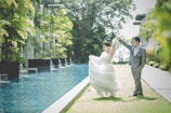 A couple is dancing gracefully next to a long, elegant swimming pool surrounded by lush greenery. The woman wears a flowing white dress, and the man is in a sleek gray suit. The setting is vibrant and serene, with tall plants and modern buildings in the background.