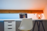 A modern workspace setup with a white desk and chair. On the desk, there is an open laptop displaying a music-related interface. To the right, an aesthetically pleasing lamp with an exposed bulb provides warm lighting. The wall is painted white with a wooden beam adding a rustic touch, and the light effect creates a gradient from blue to orange.