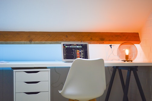 A modern workspace setup with a white desk and chair. On the desk, there is an open laptop displaying a music-related interface. To the right, an aesthetically pleasing lamp with an exposed bulb provides warm lighting. The wall is painted white with a wooden beam adding a rustic touch, and the light effect creates a gradient from blue to orange.