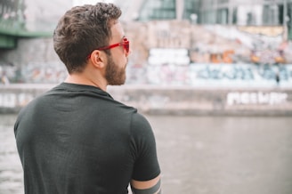 A person with short, dark hair and a beard is wearing a black t-shirt and red sunglasses. The individual is standing by a river with an urban background that features graffiti-covered walls.