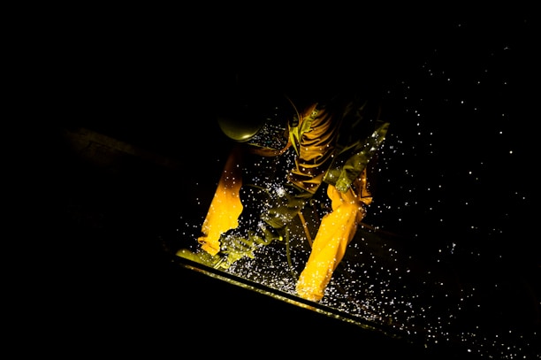A person in a dark environment is engaged in welding, wearing protective clothing and a helmet. Bright sparks fly around, illuminating parts of the person and the workspace.