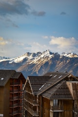 A breathtaking view of snow-capped mountains in the background under a partly cloudy sky. In the foreground, modern wooden chalets with dark roofs and multiple balconies add a cozy alpine touch.