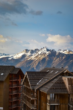 A breathtaking view of snow-capped mountains in the background under a partly cloudy sky. In the foreground, modern wooden chalets with dark roofs and multiple balconies add a cozy alpine touch.