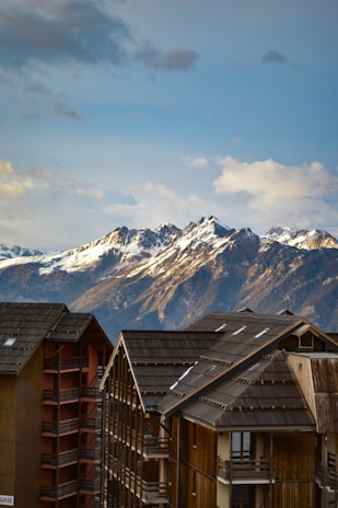 A breathtaking view of snow-capped mountains in the background under a partly cloudy sky. In the foreground, modern wooden chalets with dark roofs and multiple balconies add a cozy alpine touch.