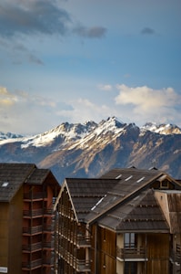 A breathtaking view of snow-capped mountains in the background under a partly cloudy sky. In the foreground, modern wooden chalets with dark roofs and multiple balconies add a cozy alpine touch.