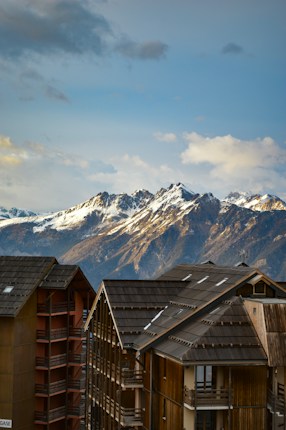 A breathtaking view of snow-capped mountains in the background under a partly cloudy sky. In the foreground, modern wooden chalets with dark roofs and multiple balconies add a cozy alpine touch.
