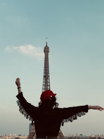 A person stands with arms outstretched, facing the Eiffel Tower under a clear blue sky, wearing a red beret and a fringed jacket.