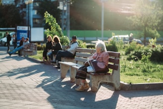 A serene park setting where an elderly person sits alone on a wooden bench, bathed in warm sunlight. In the background, a group of people is engaged in conversation on another bench. Lush greenery and a paved walkway are visible, with gentle sunlight casting long shadows.