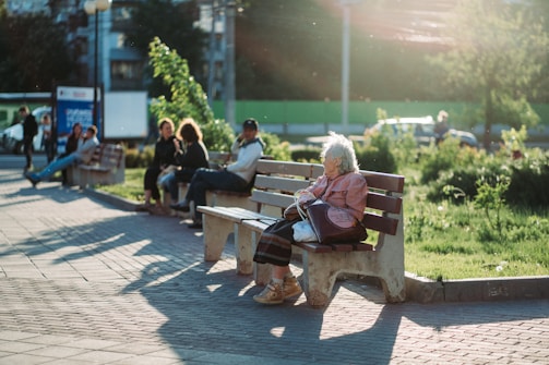 A serene park setting where an elderly person sits alone on a wooden bench, bathed in warm sunlight. In the background, a group of people is engaged in conversation on another bench. Lush greenery and a paved walkway are visible, with gentle sunlight casting long shadows.