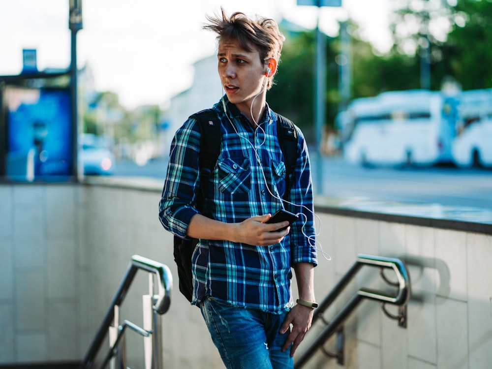 man in blue plaid dress shirt and blue distressed denim jeans with pair of white earbuds connected to smartphone in hand standing on top of subway stairs