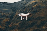 A white drone hovers in front of a mountainous landscape with a rugged terrain and a mix of dark green and brown hues.