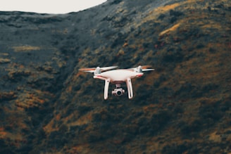 A white drone hovers in front of a mountainous landscape with a rugged terrain and a mix of dark green and brown hues.
