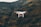 A white drone hovers in front of a mountainous landscape with a rugged terrain and a mix of dark green and brown hues.