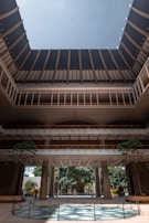 A large, open architectural structure with multiple levels and a circular opening at the top that allows in natural light. The building features a modern design with tall pillars and a spacious courtyard area. Lush greenery and trees can be seen inside and surrounding the structure, adding a natural element to the setting.