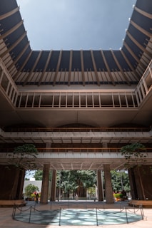 A large, open architectural structure with multiple levels and a circular opening at the top that allows in natural light. The building features a modern design with tall pillars and a spacious courtyard area. Lush greenery and trees can be seen inside and surrounding the structure, adding a natural element to the setting.
