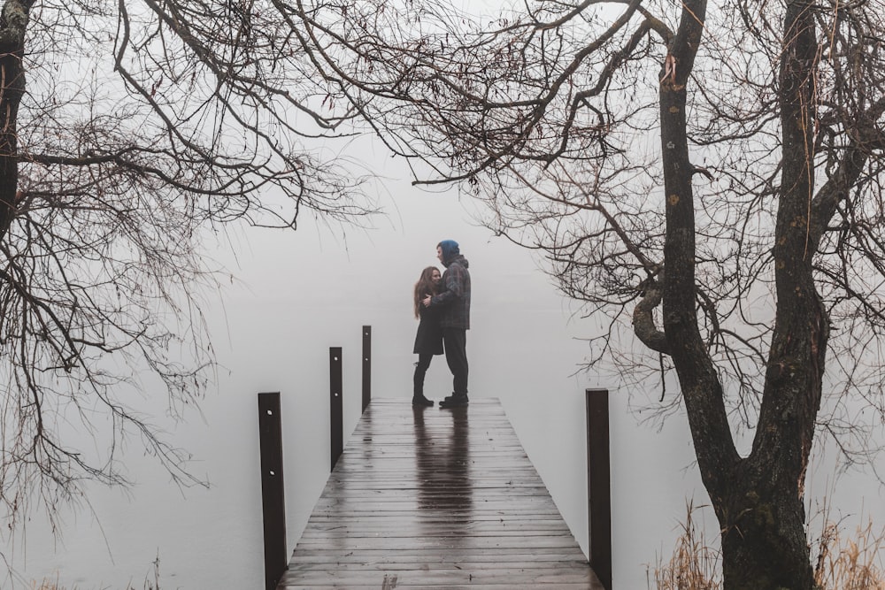 man and woman facing each other and standing on beach dock surrounded with bare trees under foggy season