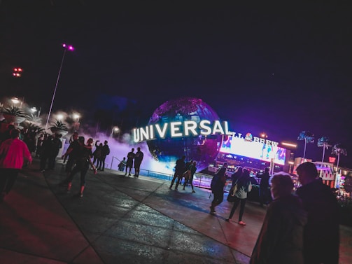 A large illuminated globe with the word 'Universal' is prominent at the center, surrounded by a misty, atmospheric effect. People are walking around the area, with some standing and looking at the globe. In the background, there are bright electronic billboards and palm trees, contributing to a lively and bustling environment.