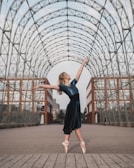 A ballerina in a blue dress stands en pointe, arms outstretched gracefully under an arched, open metal framework. The background includes urban architectural elements like brick and glass buildings, adding a modern feel to the scene.