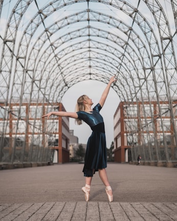 A ballerina in a blue dress stands en pointe, arms outstretched gracefully under an arched, open metal framework. The background includes urban architectural elements like brick and glass buildings, adding a modern feel to the scene.