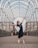 A ballerina in a blue dress stands en pointe, arms outstretched gracefully under an arched, open metal framework. The background includes urban architectural elements like brick and glass buildings, adding a modern feel to the scene.