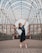 A ballerina in a blue dress stands en pointe, arms outstretched gracefully under an arched, open metal framework. The background includes urban architectural elements like brick and glass buildings, adding a modern feel to the scene.