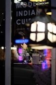 A person sitting at a table inside a restaurant, viewed through a window with reflections of neon lights. On the window, the words 'Modern Indian Cuisine' along with location names like 'London' and 'New' are visible. The table is set with a small vase containing a plant, a bottle of water, a glass, and a napkin holder. Dim, ambient lighting suggests a cozy dining atmosphere.