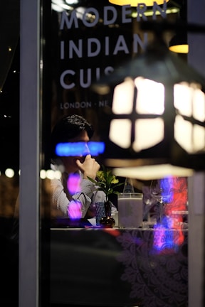 A person sitting at a table inside a restaurant, viewed through a window with reflections of neon lights. On the window, the words 'Modern Indian Cuisine' along with location names like 'London' and 'New' are visible. The table is set with a small vase containing a plant, a bottle of water, a glass, and a napkin holder. Dim, ambient lighting suggests a cozy dining atmosphere.