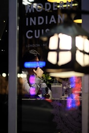 A person sitting at a table inside a restaurant, viewed through a window with reflections of neon lights. On the window, the words 'Modern Indian Cuisine' along with location names like 'London' and 'New' are visible. The table is set with a small vase containing a plant, a bottle of water, a glass, and a napkin holder. Dim, ambient lighting suggests a cozy dining atmosphere.