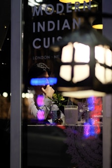 A person sitting at a table inside a restaurant, viewed through a window with reflections of neon lights. On the window, the words 'Modern Indian Cuisine' along with location names like 'London' and 'New' are visible. The table is set with a small vase containing a plant, a bottle of water, a glass, and a napkin holder. Dim, ambient lighting suggests a cozy dining atmosphere.